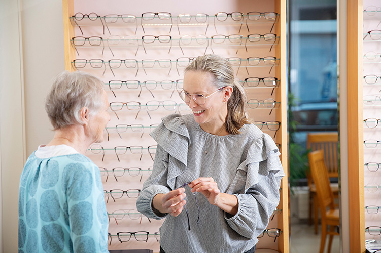 Optikko esittelee asiakkaalle sopivia silmälasikehyksiä kehyshyllyn edessä. Myymälässä on rauhallinen ilmapiiri.